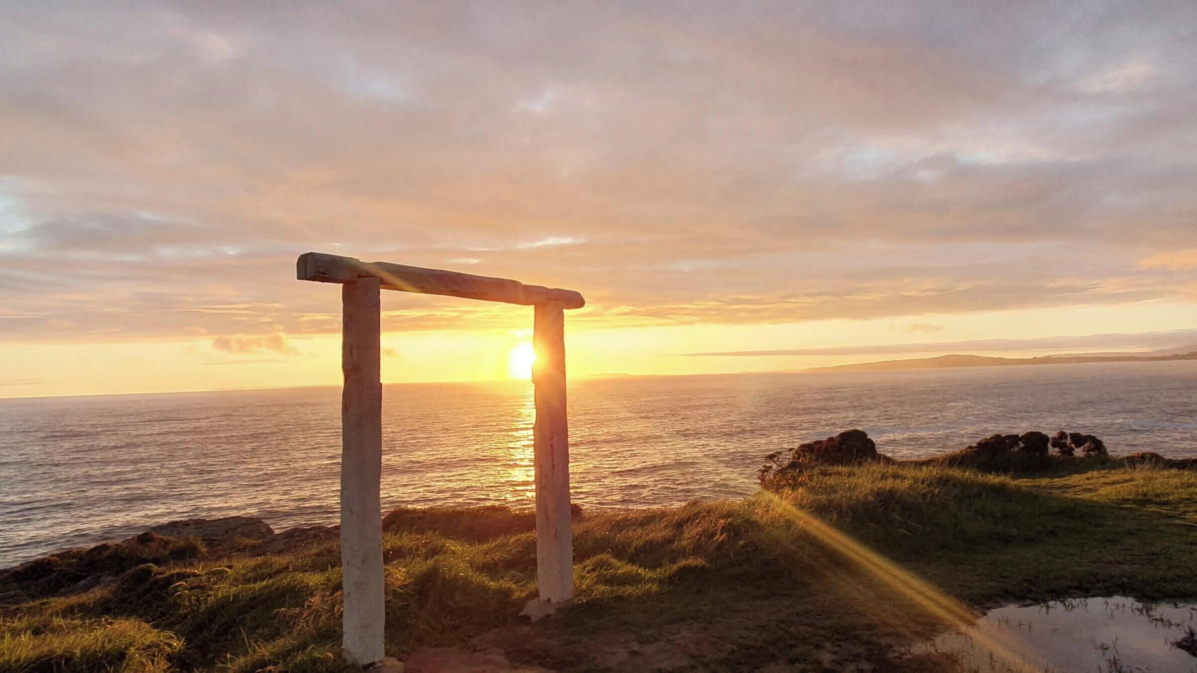 Une structure en pierre avec une poutre horizontale, surplombant une étendue d’eau, avec un coucher de soleil en arrière-plan.