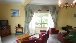 Una habitación con una mesa de madera, sillas rojas, un televisor y un amplio ventanal con cortinas.