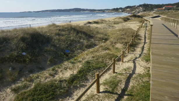 A wooden boardwalk along a beach with a clear view of the ocean and distant hills. - Hotel La Costera (Sanxenxo)