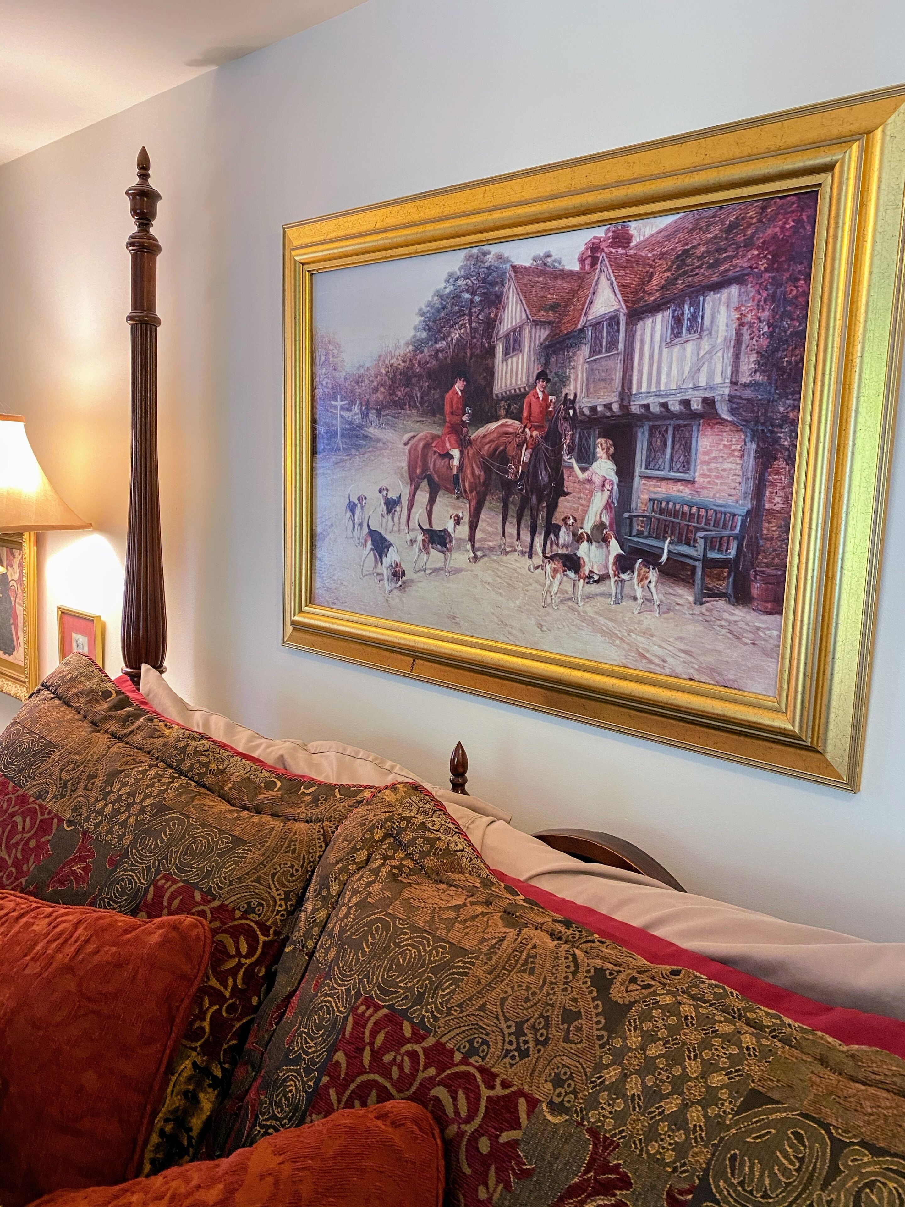 Una habitación con un cuadro grande enmarco de una escena histórica, una cama de madera con cabecera tallada y un plaid estampado en el sofá.