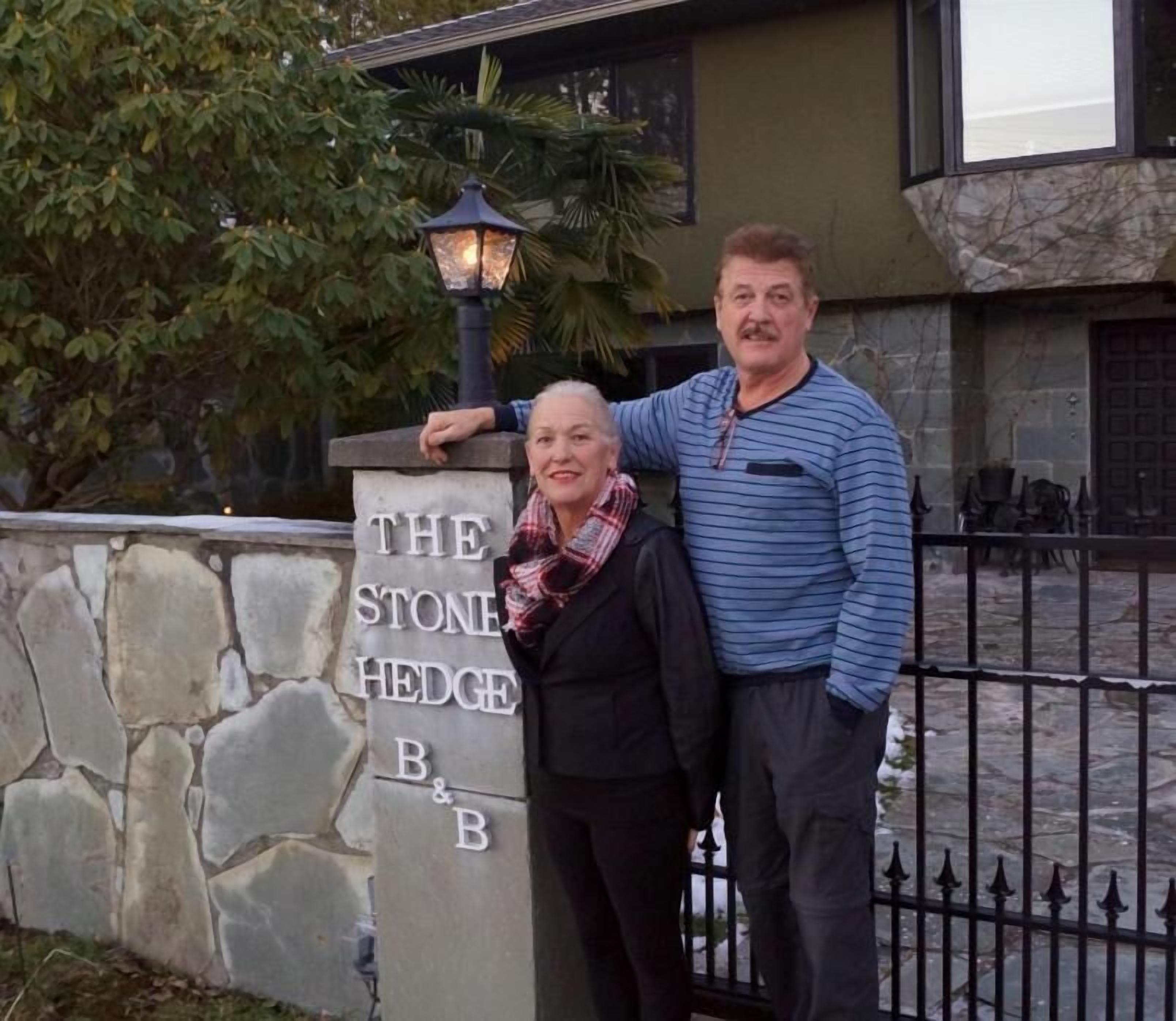 Two people standing in front of a stone pillar with the inscription "THE STONE HEDGE B&B".