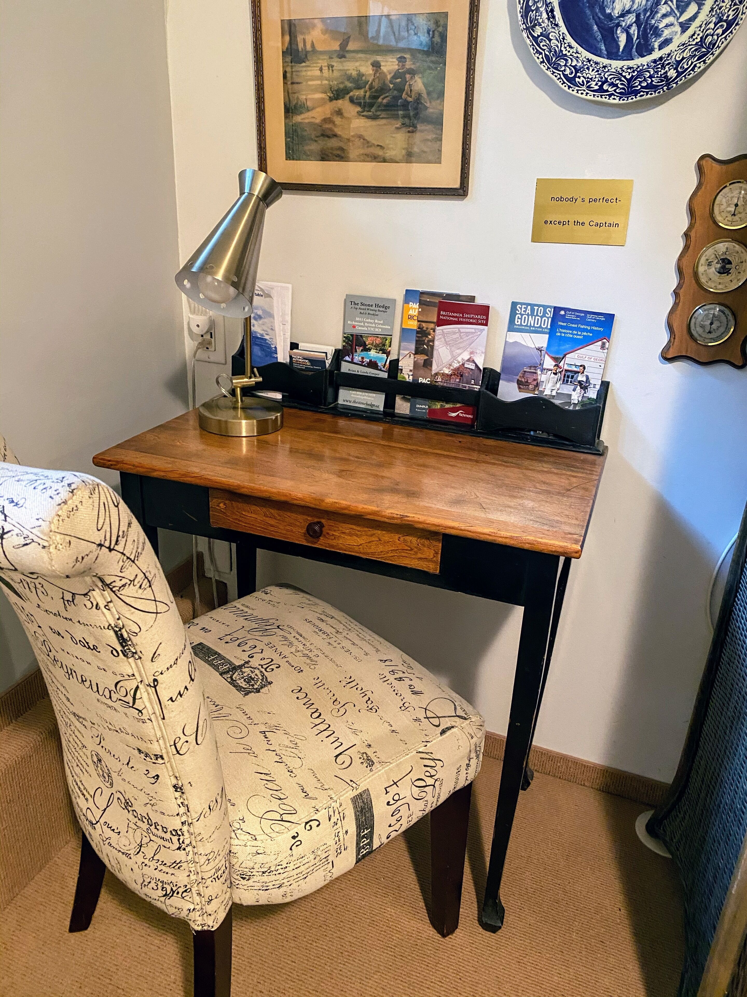 Un escritorio de madera con una lámpara, una silla con un cojín estampado, un cuadro enmarcado y un reloj en la pared.