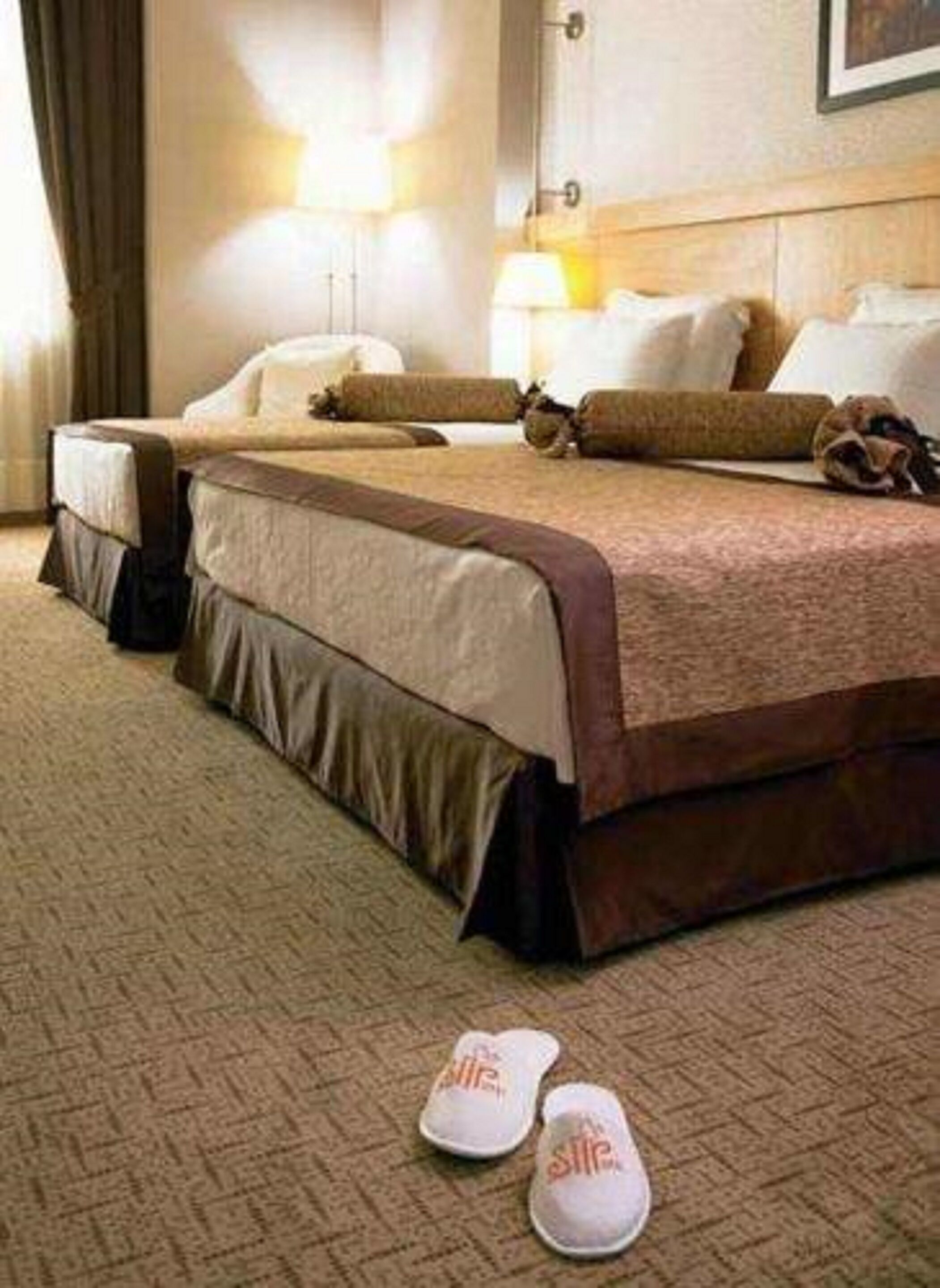 A hotel room with two beds, a carpeted floor, and a pair of slippers on the floor.