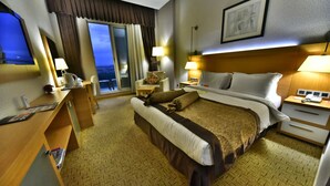 Een hotelkamer met een groot bed, een bureau met een computer, een stoel, een nachtkastje met een lamp, en een raam met uitzicht.
