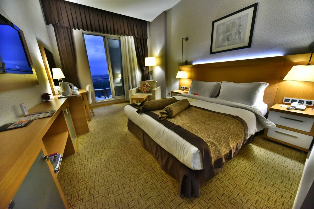 A hotel room with a large bed, a desk with a computer, a chair, a nightstand with a lamp, and a window with a view.