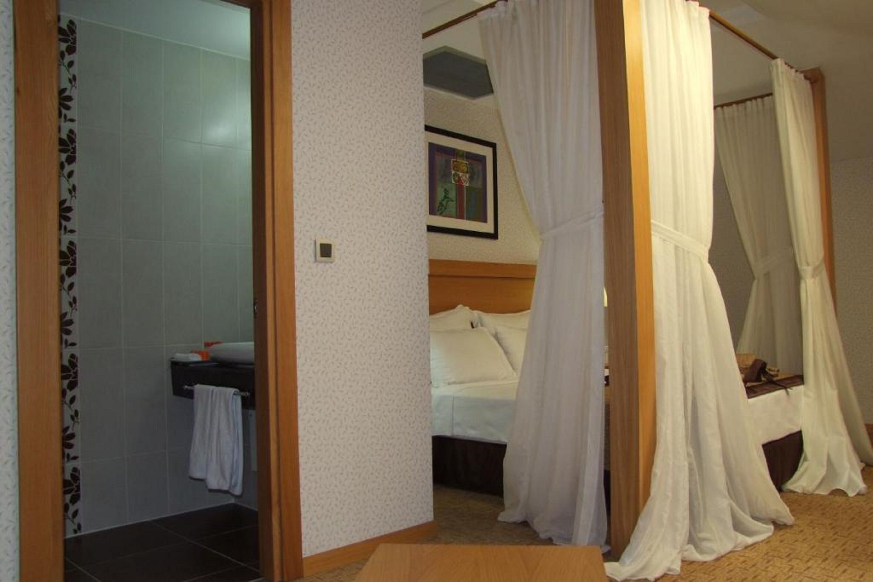 Un dormitorio con cama, cortinas blancas, cabecera, un cuadro en la pared y un baño con una toalla colgada en la puerta.