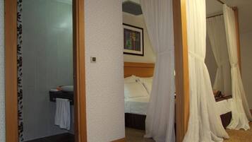 Un dormitorio con cama, cortinas blancas, cabecera, un cuadro en la pared y un baño con una toalla colgada en la puerta.