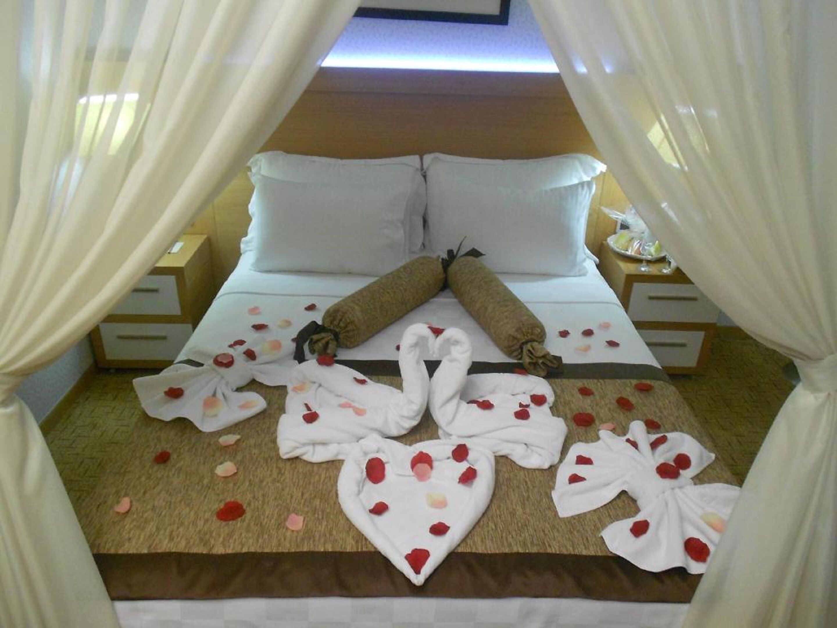 A neatly made bed with white linens, two rolled towels, and rose petals scattered on the bed and floor.