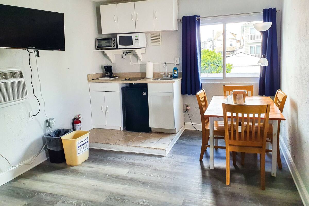 Een compacte keuken met witte kastjes, een magnetron en een koffiezetapparaat. Er is een eettafel met stoelen en een raam met gordijnen.