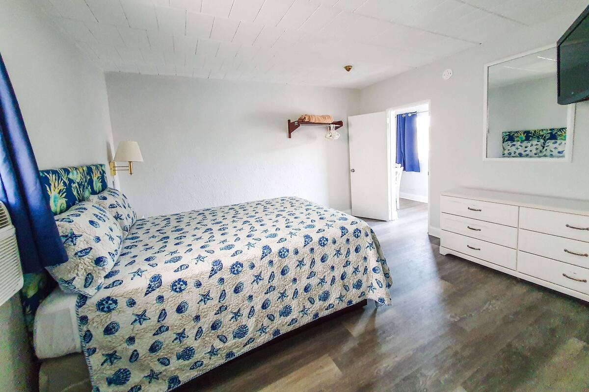 Een slaapkamer met een bed, een dressoir, een spiegel en een deur naar een andere kamer.
