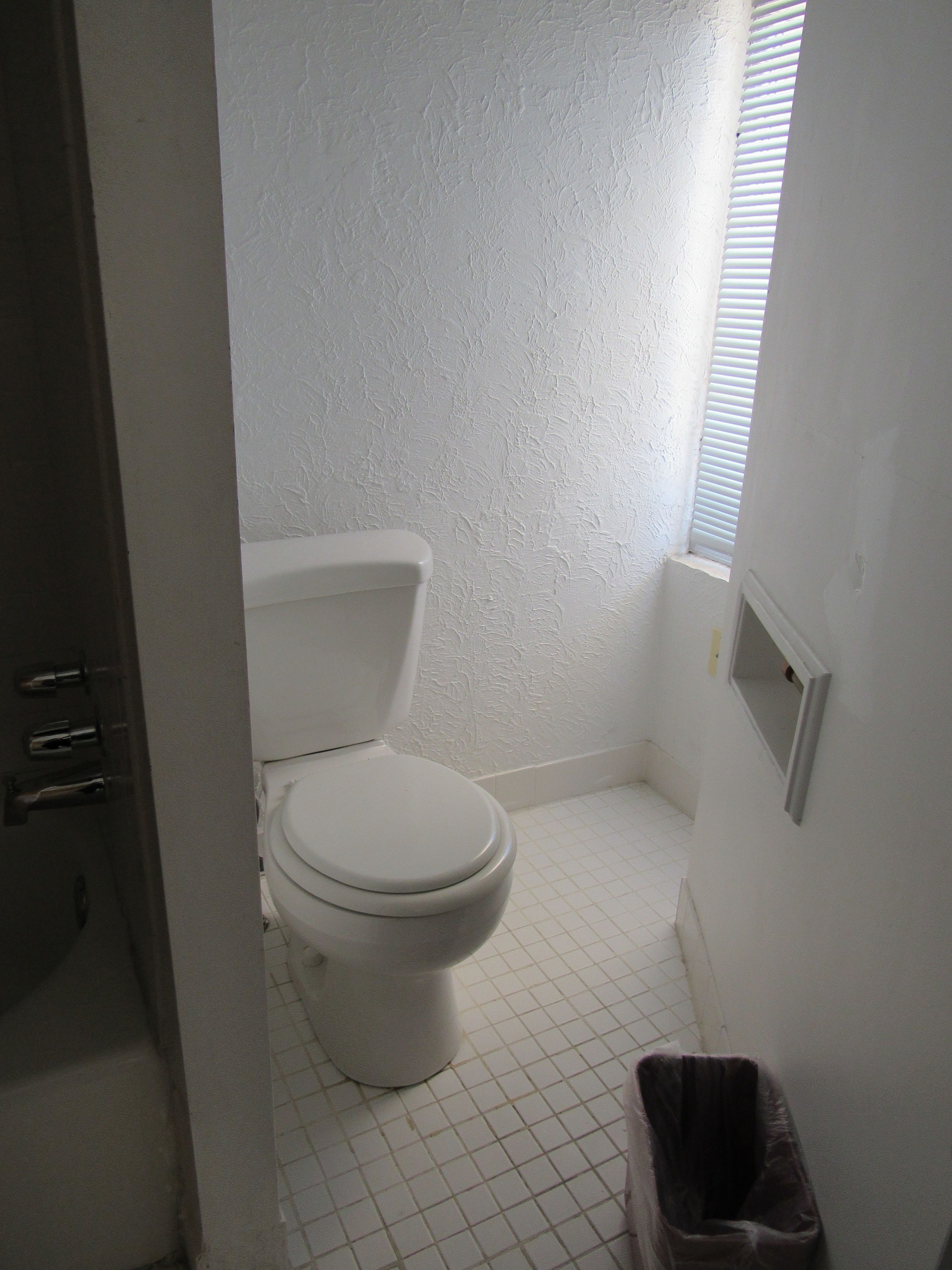 Une petite salle de bain blanche, avec des toilettes, une poubelle et une fenêtre.