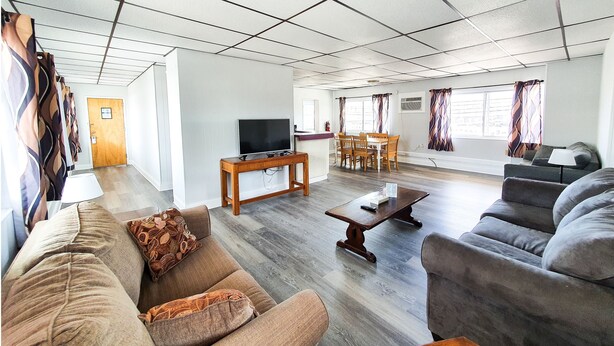 Amplia sala de estar con sofá, sillón y mesa de centro. Hay un televisor sobre un mueble bajo, una zona de comedor con sillas y una ventana con cortinas.