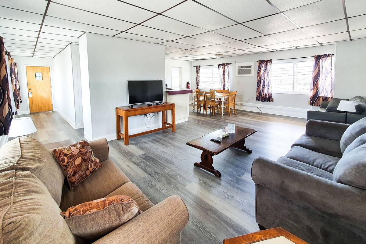 Een ruime woonkamer met een bank, fauteuil en salontafel. Er is een televisie op een dressoir, een eethoek met stoelen, en een raam met gordijnen.