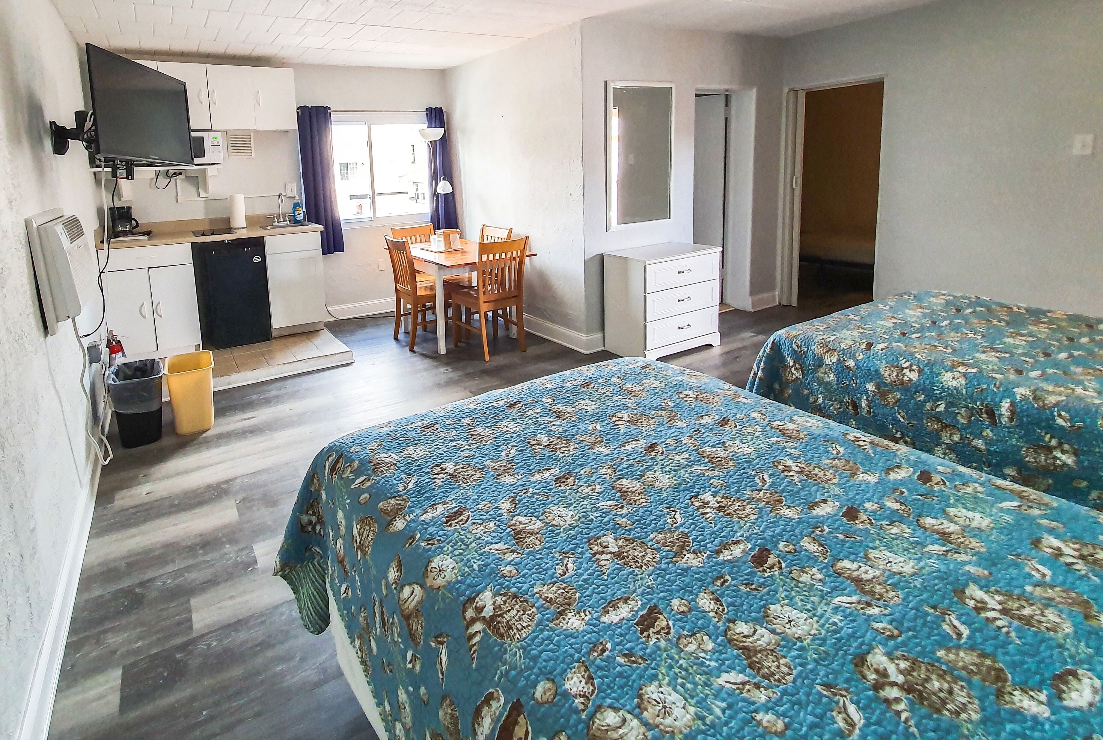 A compact hotel room with two beds, a kitchenette, a dining area, and a bathroom.