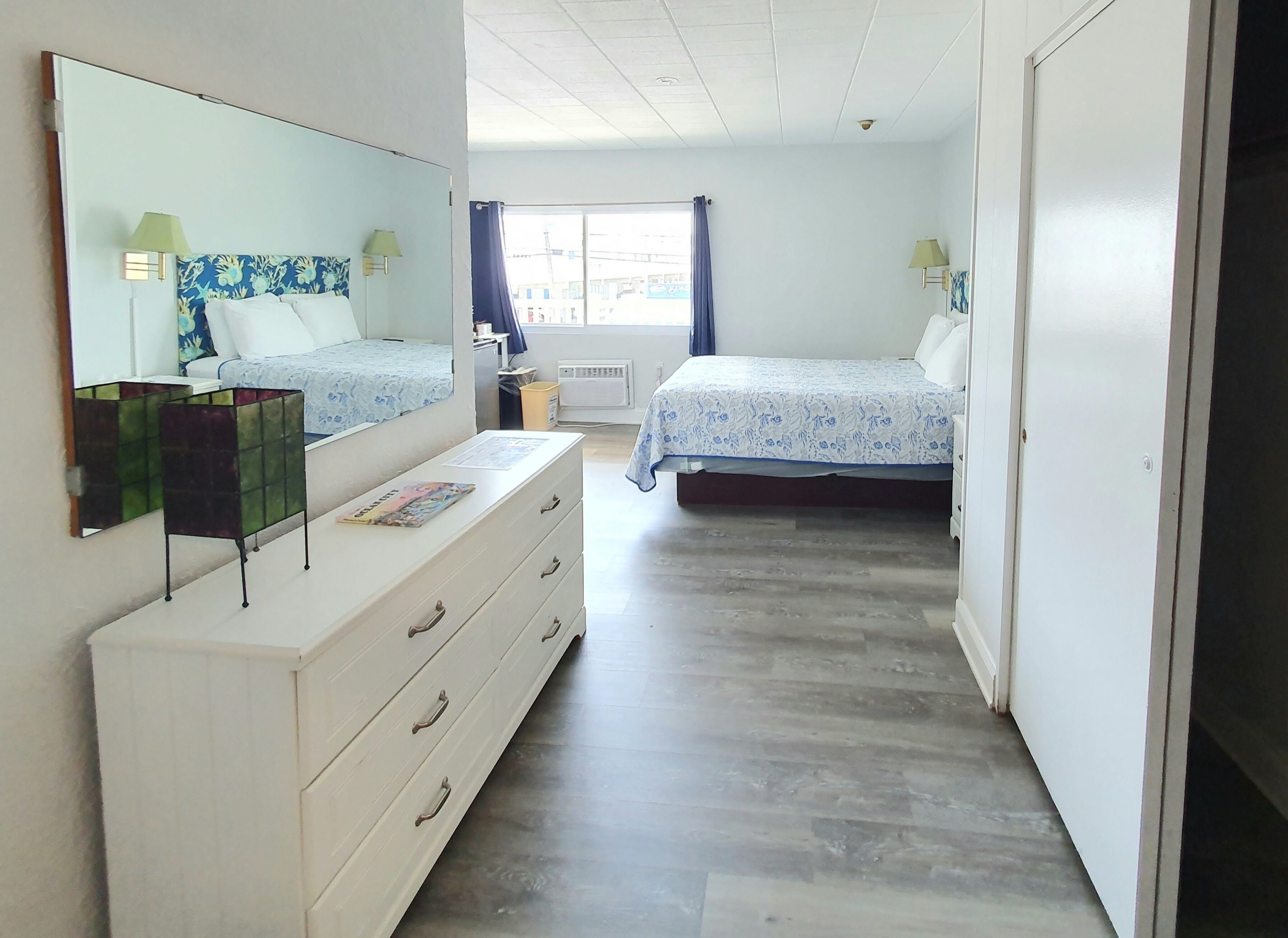 Une chambre avec un grand miroir, une commode, un lit recouvert d’une couvre-lit à motifs floraux et une fenêtre avec des rideaux.