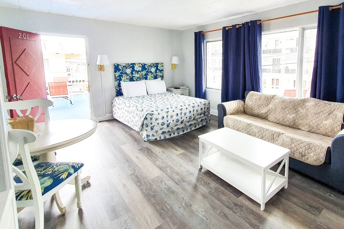 Een kamer met een bed, een bank, een klein tafeltje en een stoel. Er is een raam met gordijnen en een lamp aan de muur.