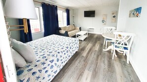 Un dormitorio con cama, escritorio y silla. Hay una ventana con cortinas, un televisor de pantalla plana en la pared y una obra de arte en la pared.