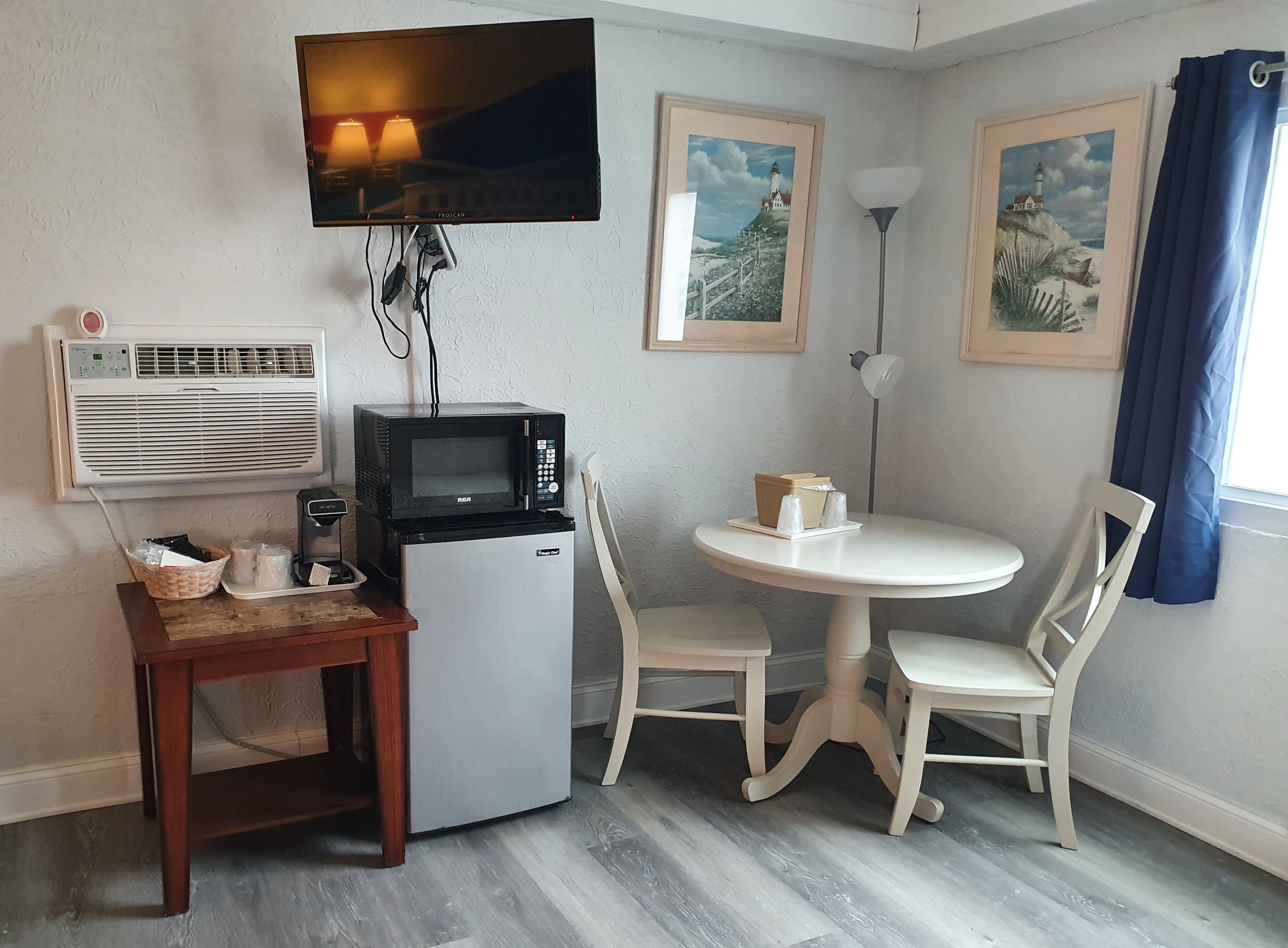 A compact kitchenette with a microwave, a small refrigerator, a table with chairs, a television mounted on the wall, and framed pictures on the wall.