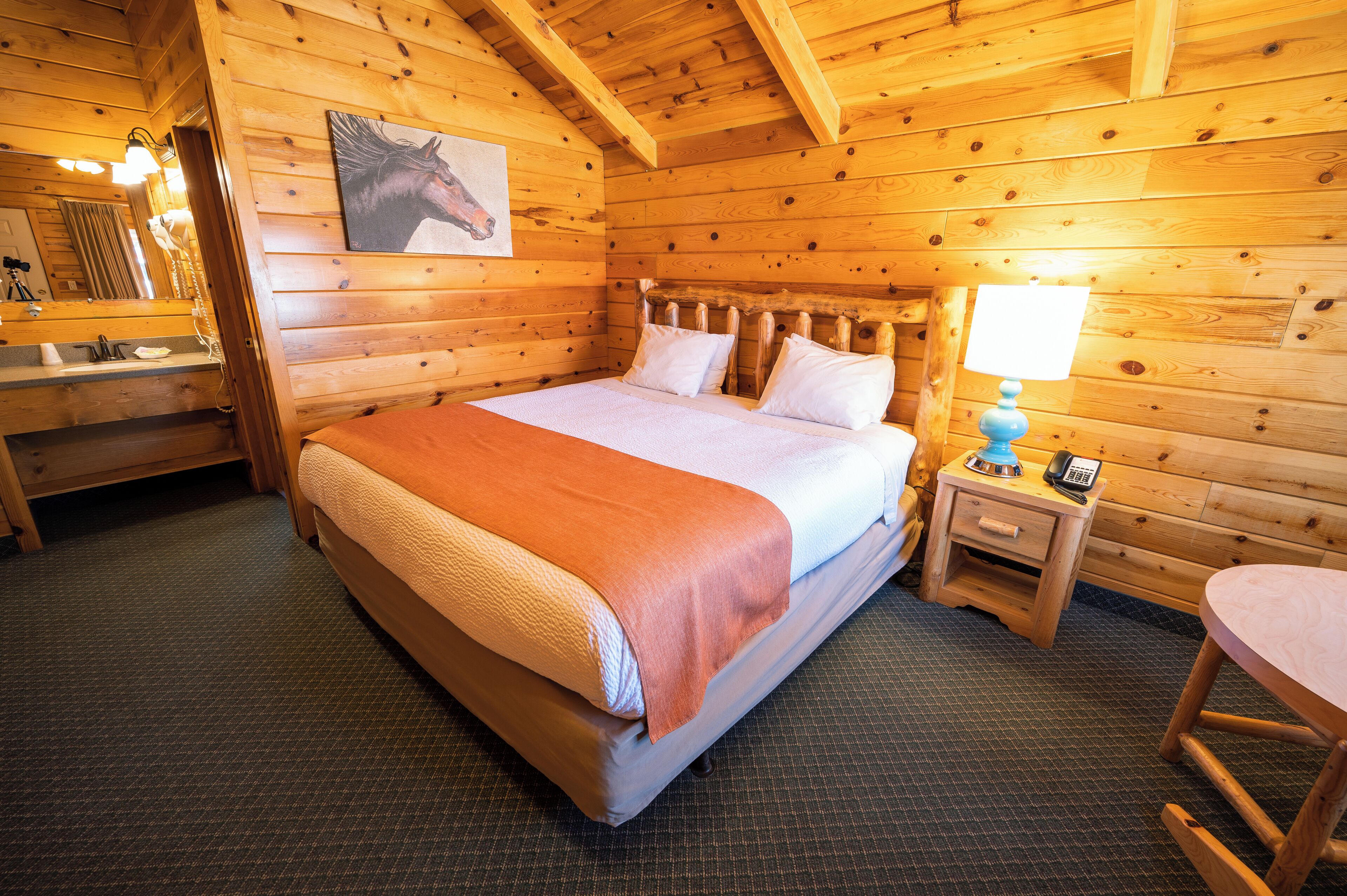 A wooden cabin room with a large bed, a bedside table with a lamp, and a small chair.
