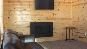 A log cabin interior with a leather sofa, a flat-screen TV mounted on the wall, and a fireplace below.