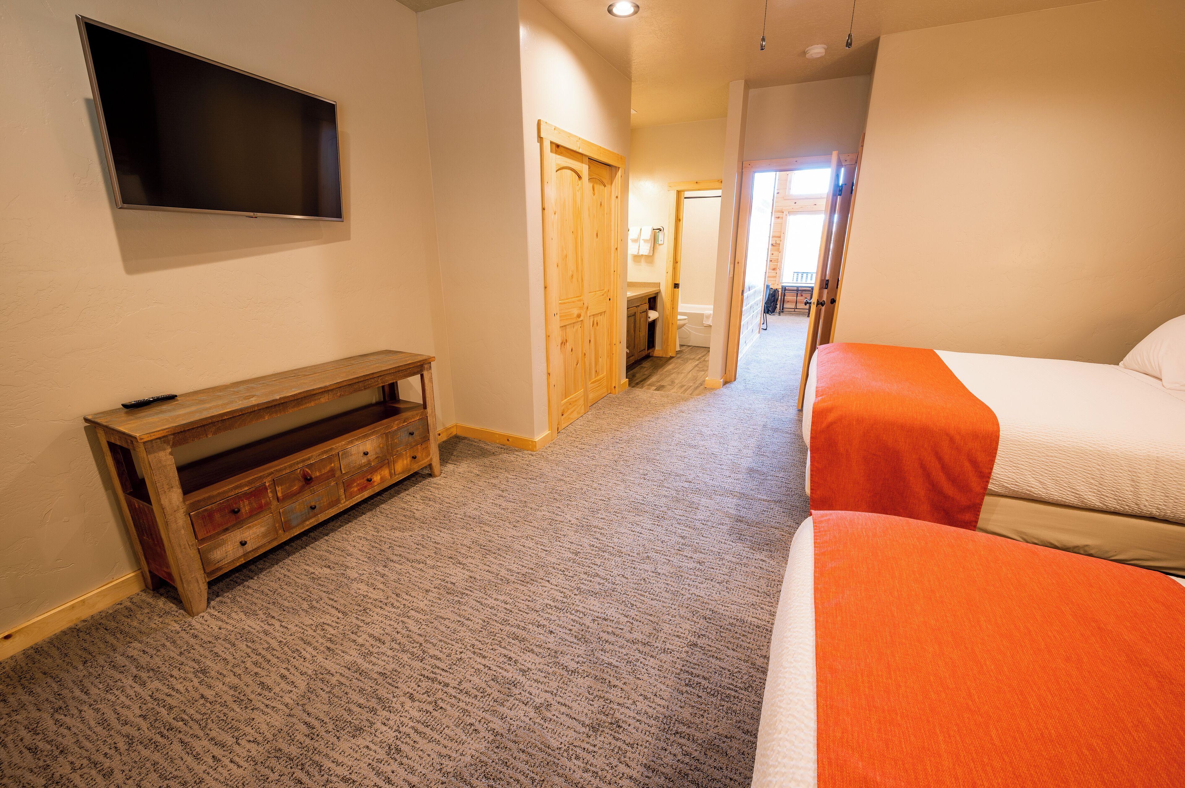 A hotel room with a TV mounted on the wall, a wooden bench, and two beds with orange bedding.