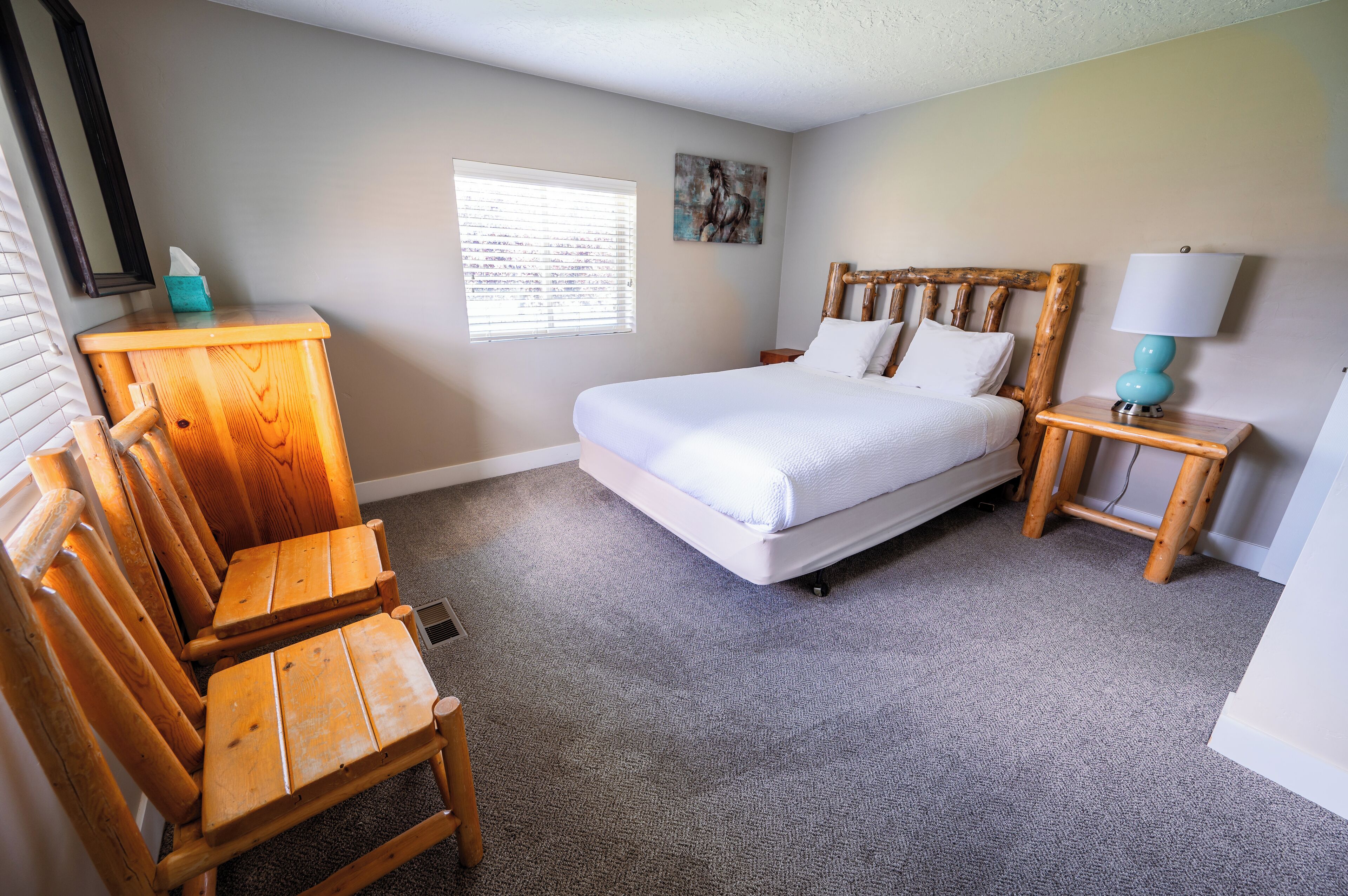 Une chambre avec un lit en bois, une table de chevet avec une lampe, une chaise en bois, une fenêtre avec des stores et un tableau accroché au mur.