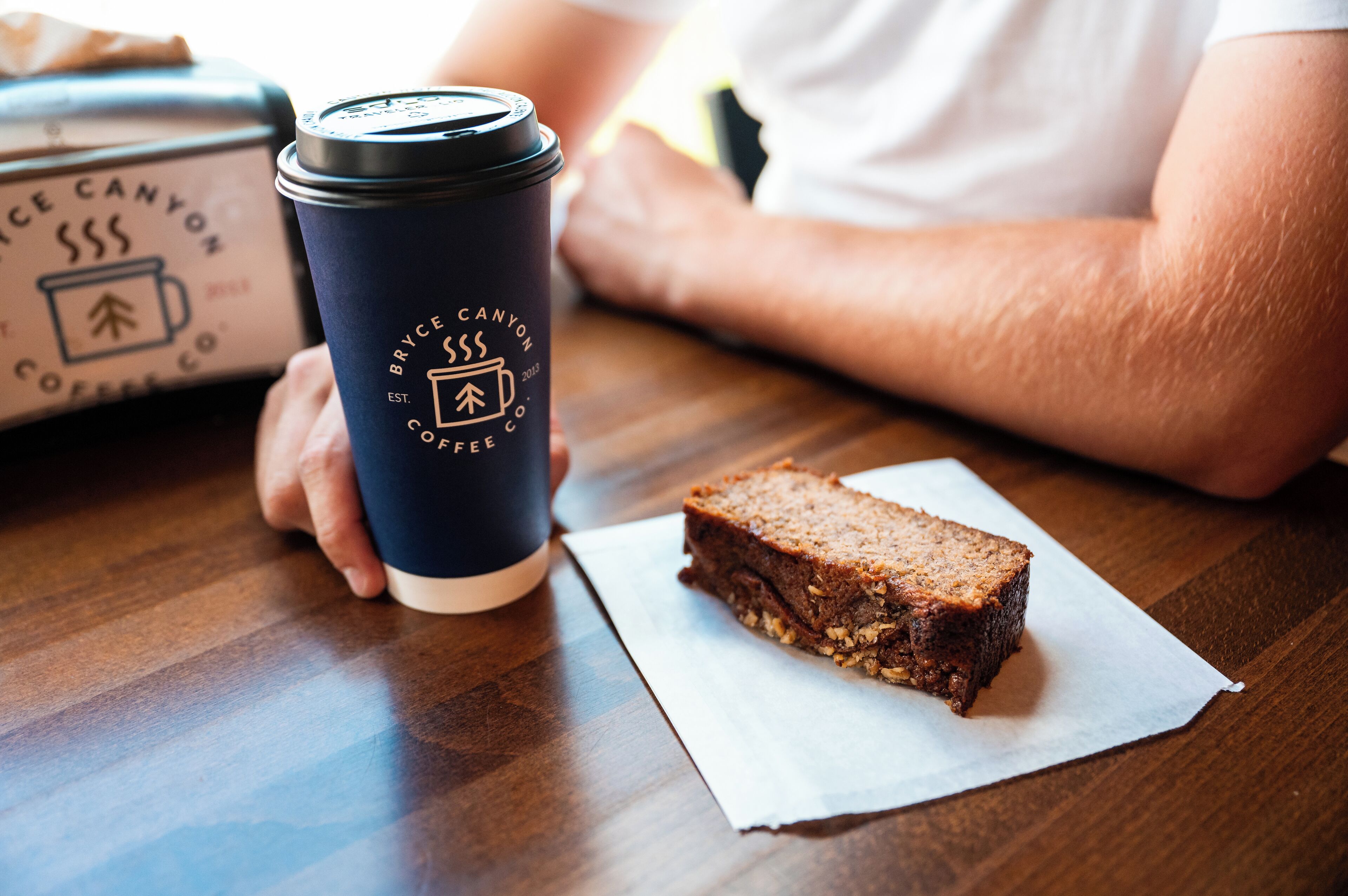 A dark blue coffee cup with a logo and text, a slice of cake on a napkin, and a box with a logo on a wooden table.