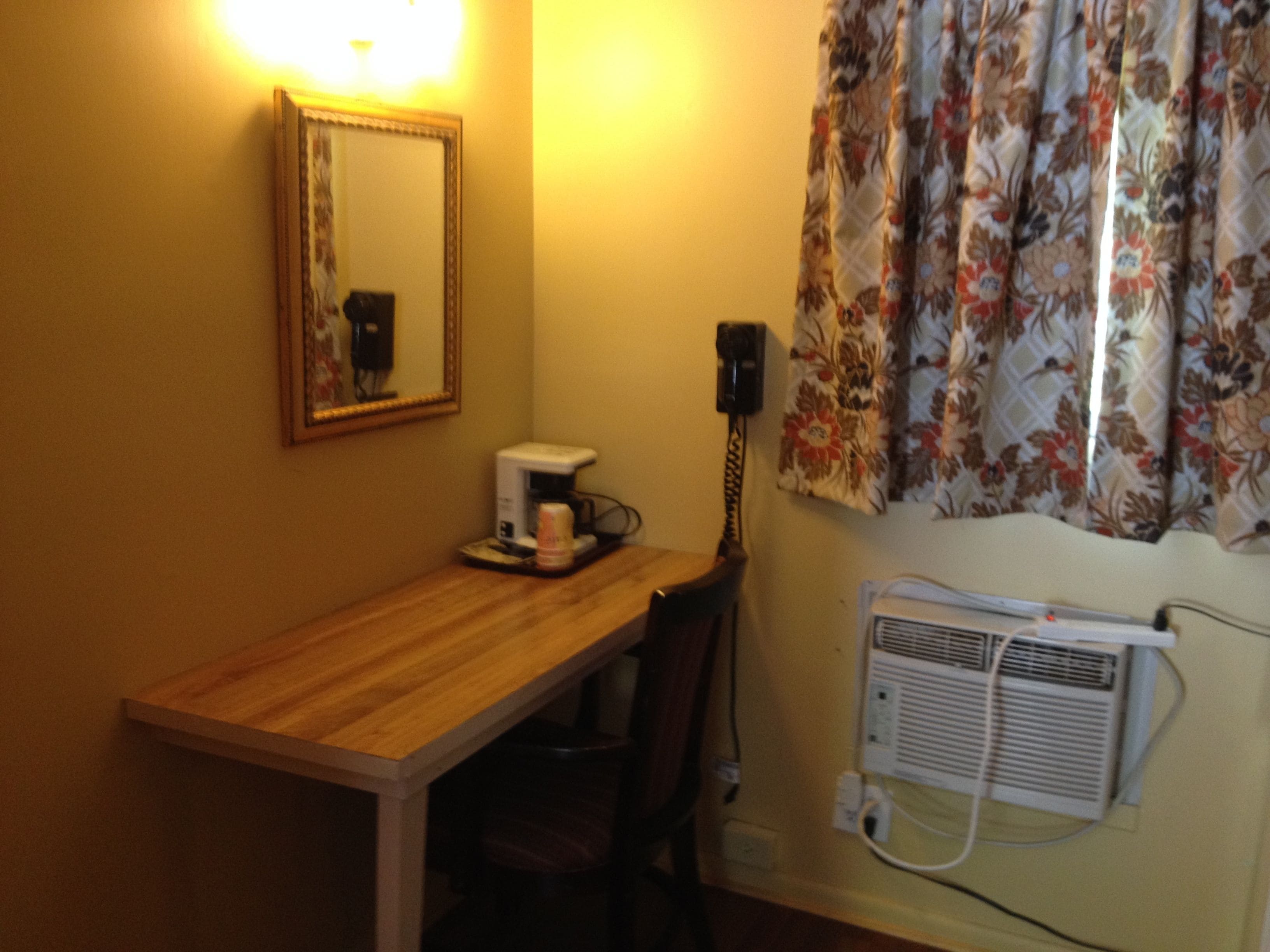 Ein kleines Zimmer mit Schreibtisch aus Holz, einer Kaffeemaschine, einem Sessel, einem Spiegel, einem Telefon und einer Wandklimaanlage.