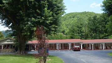 Un motel con zona de estacionamiento techada, un gran árbol en primer plano y una colina boscosa al fondo.