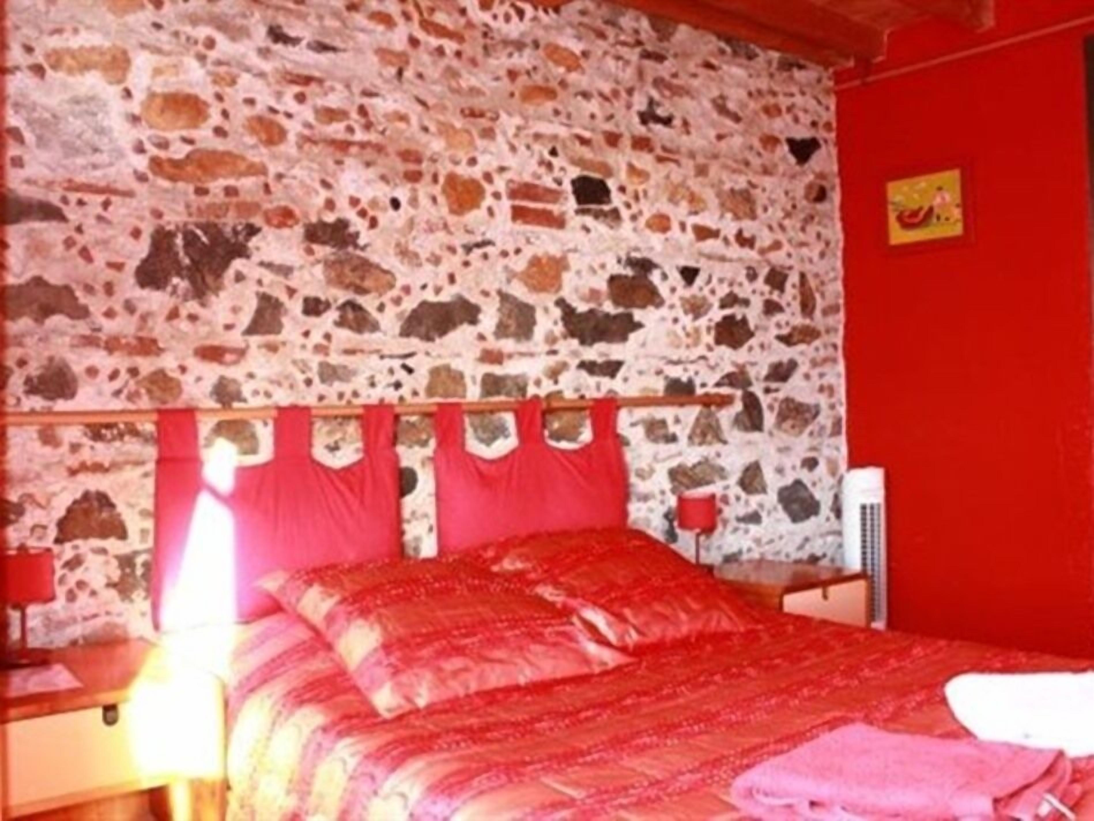 Una habitación con pared de piedra, cama con ropa de cama roja, cabecero de madera, mesita de noche con lámpara y pared roja con un pequeño cuadro.