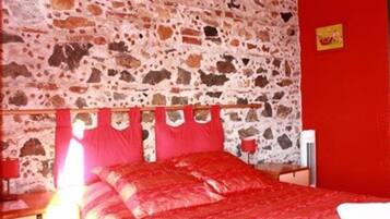 Una habitación con pared de piedra, cama con ropa de cama roja, cabecero de madera, mesita de noche con lámpara y pared roja con un pequeño cuadro.