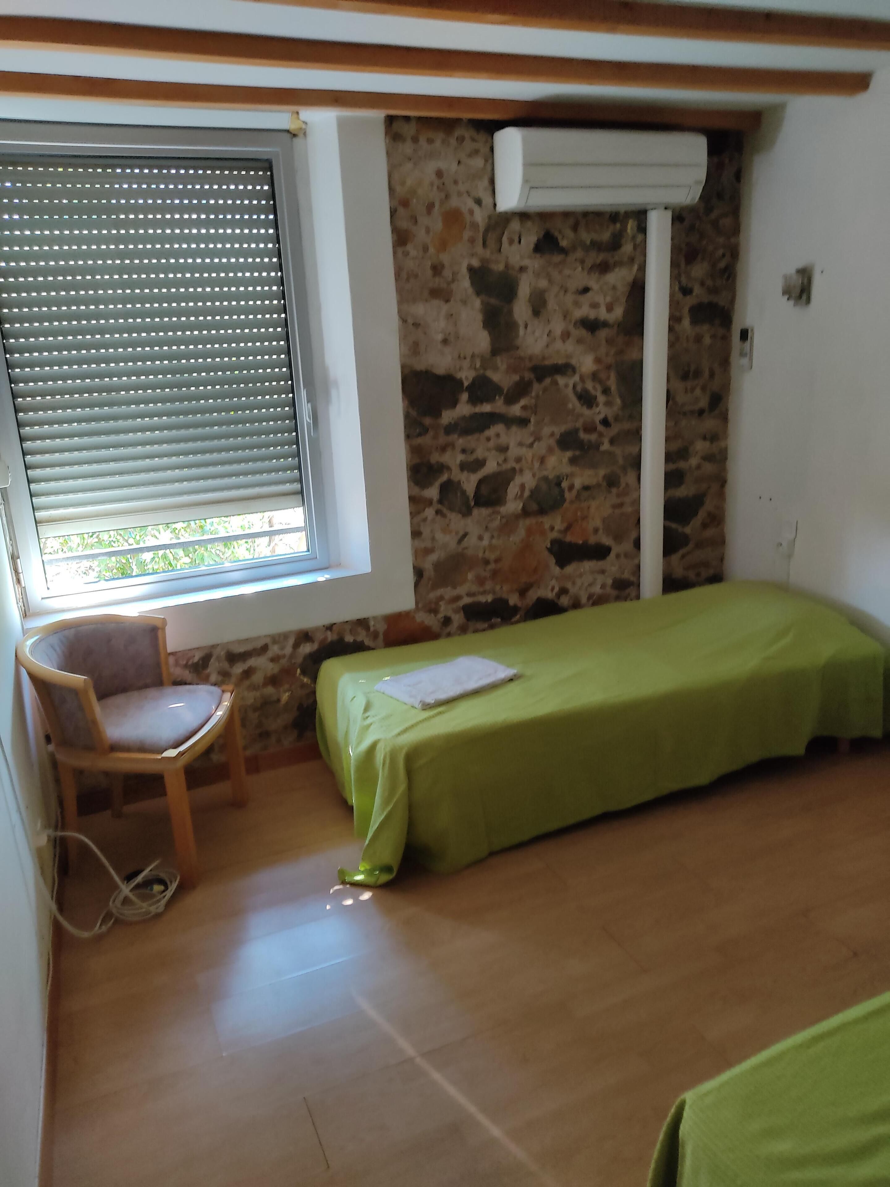 Una habitación pequeña con una cama individual, una silla, un ventanal con persianas y una pared de piedra.
