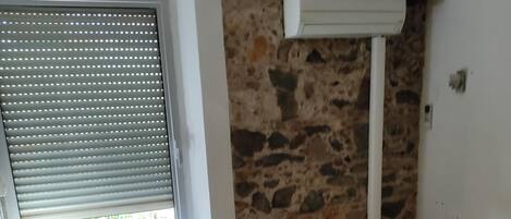 Una habitación pequeña con una cama individual, una silla, un ventanal con persianas y una pared de piedra.
