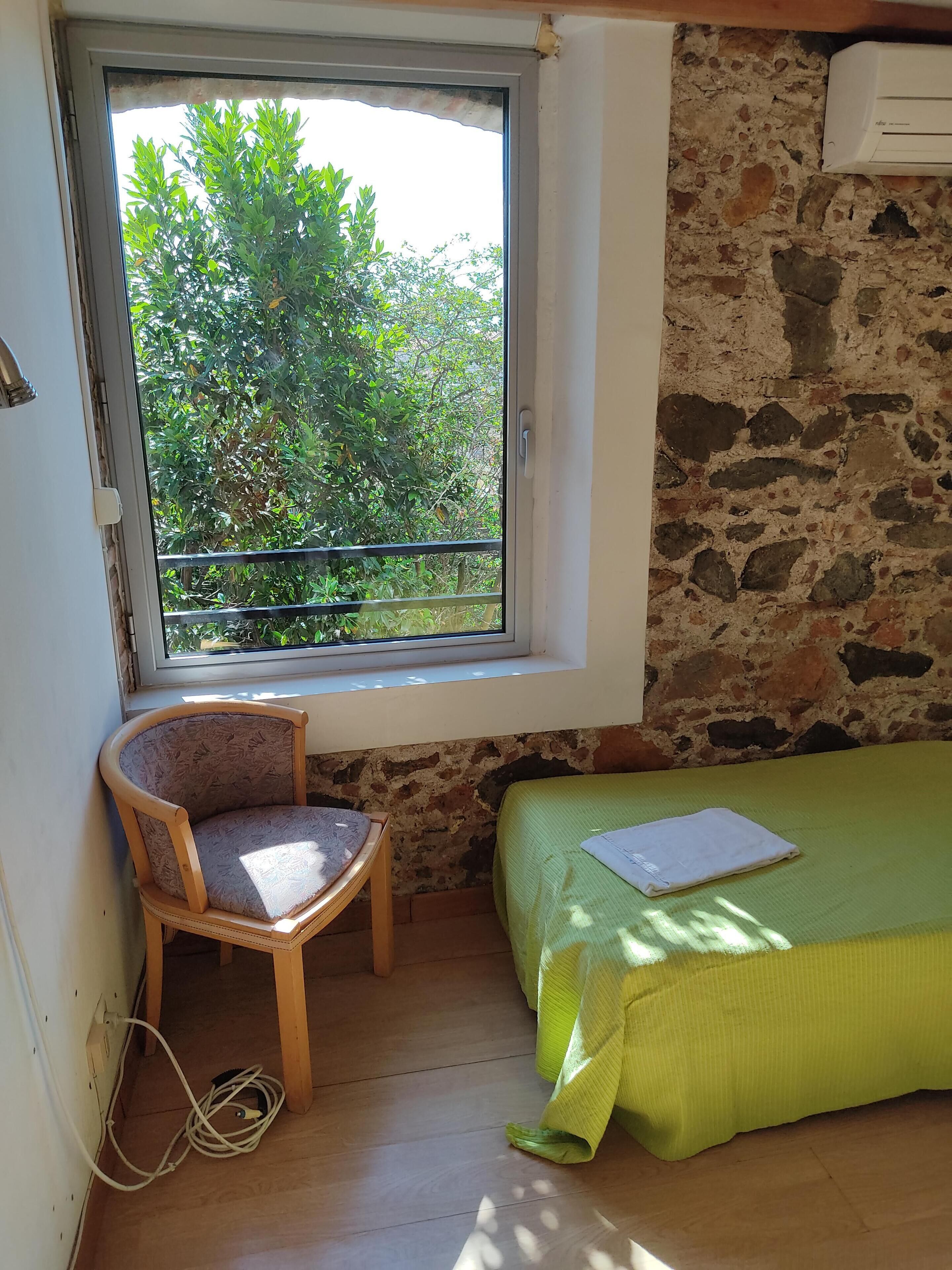 A small room with a window, a chair, a bed, and a stone wall.