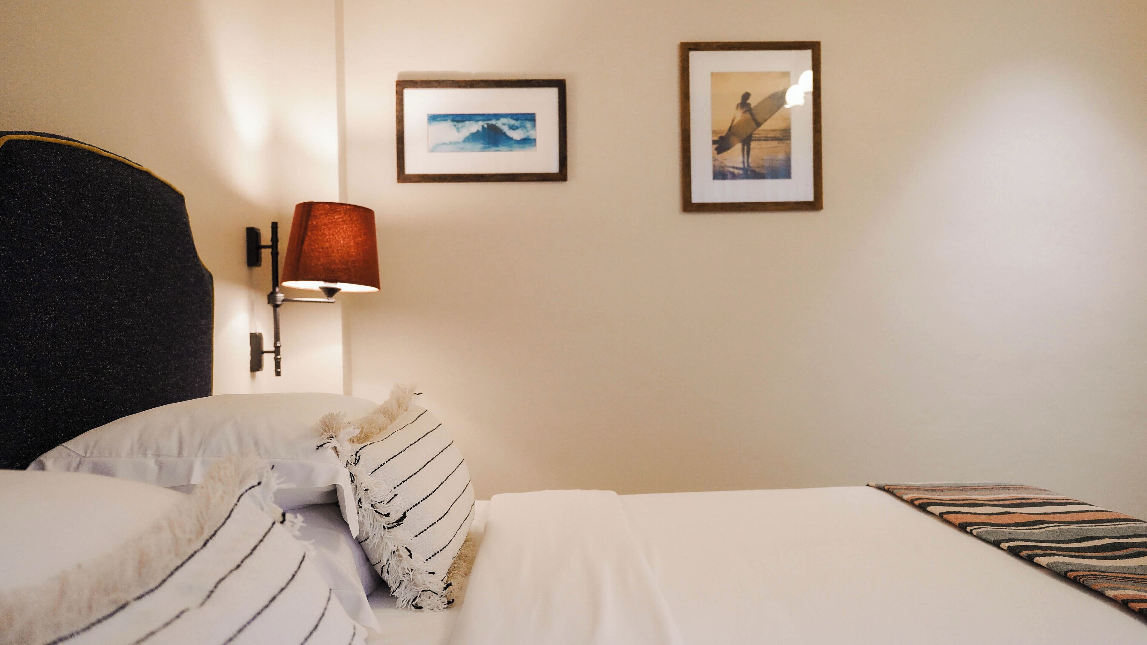 Una cama bien hecha con sábanas blancas y almohadas a rayas, un cabecero oscuro, una lámpara de pared y dos cuadros enmarcados.