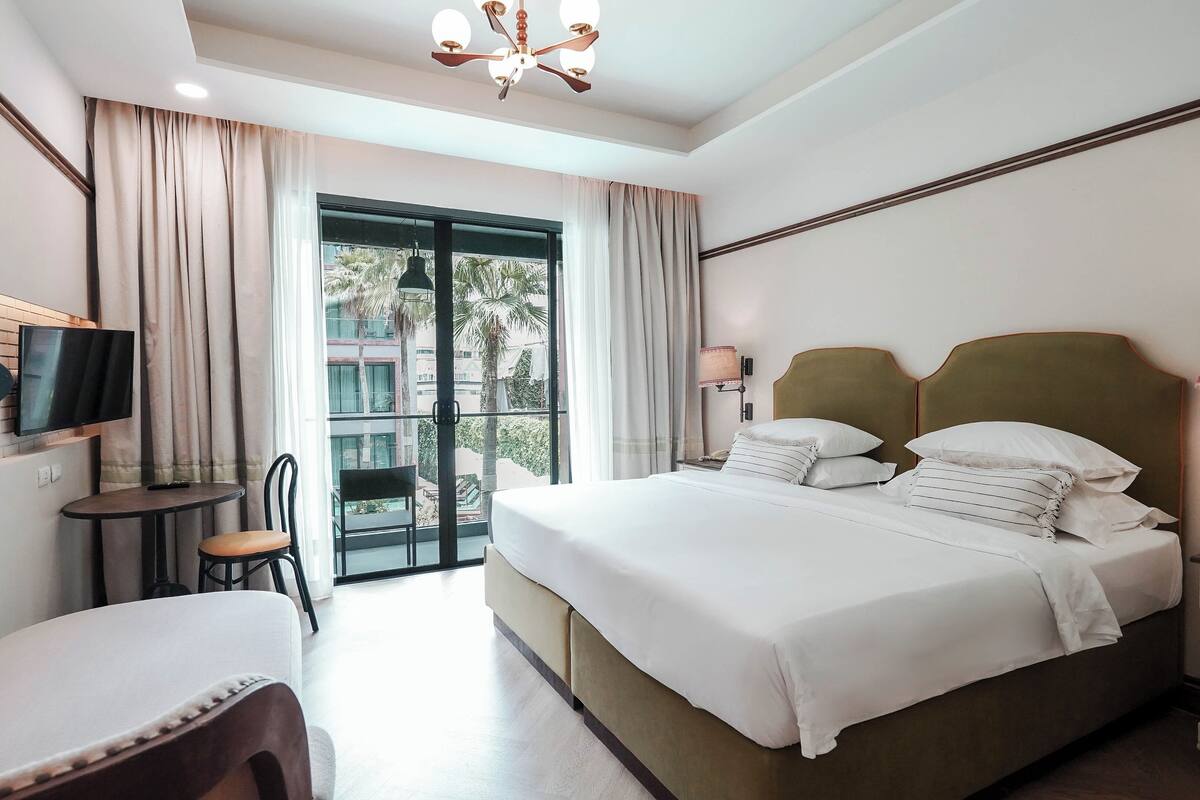 Habitación de hotel con cama, mesita de noche, lámpara, televisor y vista al exterior.