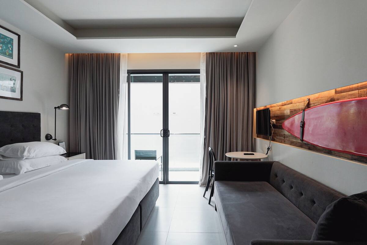Habitación de hotel con cama, zona de estar con mesa, televisor montado en un panel de madera y un ventanal con cortinas.
