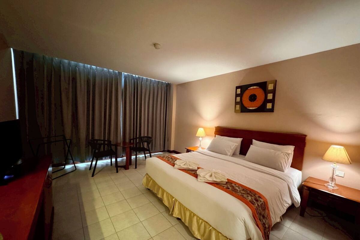 A hotel room with a large bed, two bedside tables with lamps, a TV, a desk with a chair, and a window with curtains.