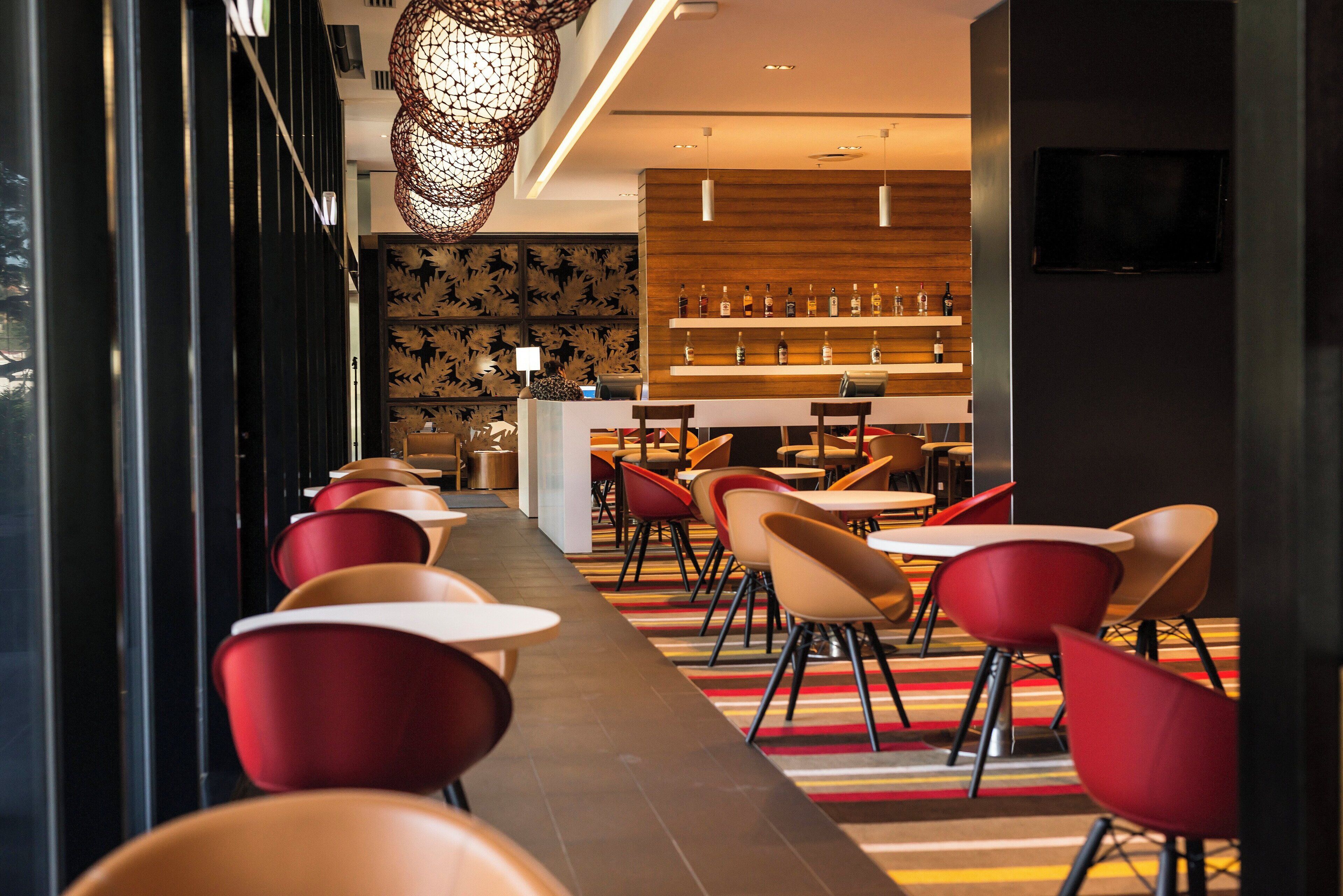 A modern hotel lobby with a long dining area, red chairs, and a bar with bottles.