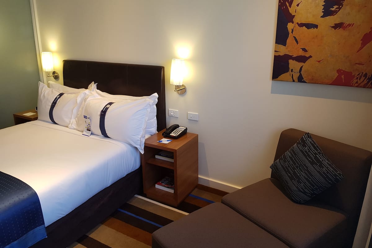 Habitación de hotel con cama, mesita de noche con teléfono, pequeño mueble auxiliar de madera, sofá y un cuadro abstracto colorido en la pared.