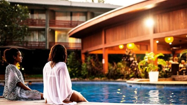 Dues persones assegudes al costat d'una piscina amb un edifici al fons.