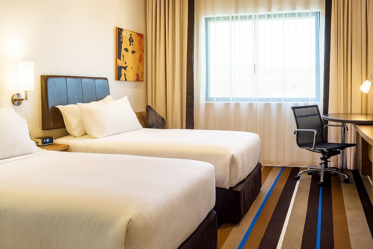A hotel room with two beds, a desk, a chair, a TV, and a painting on the wall.