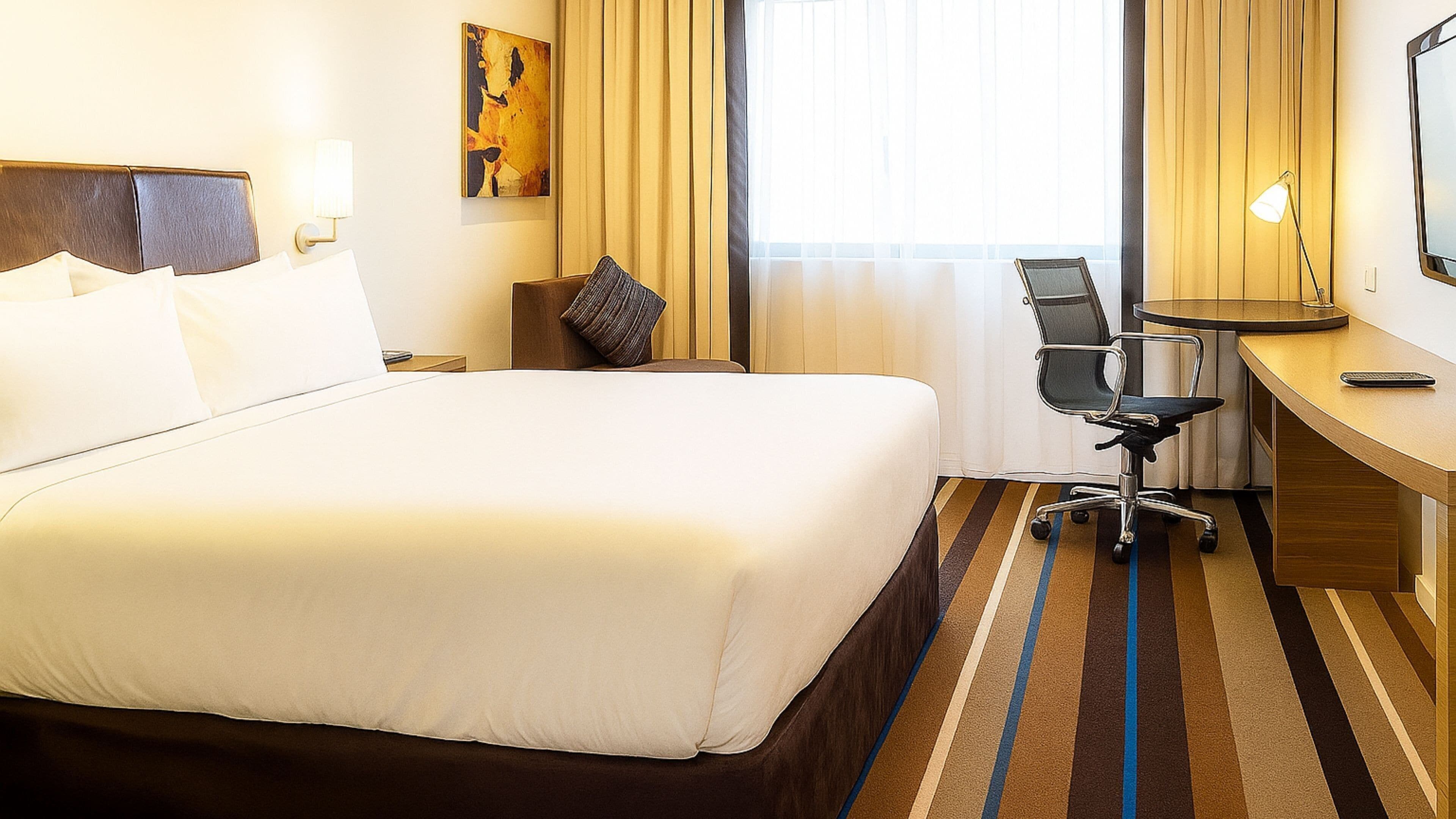 A hotel room with a large bed, a desk, a chair, and a window with curtains.