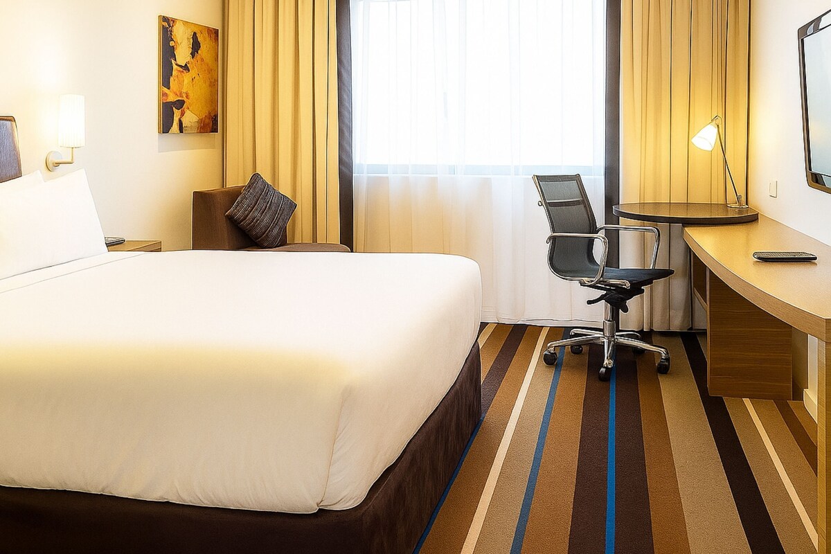 A hotel room with a large bed, a desk, a chair, and a window with curtains.