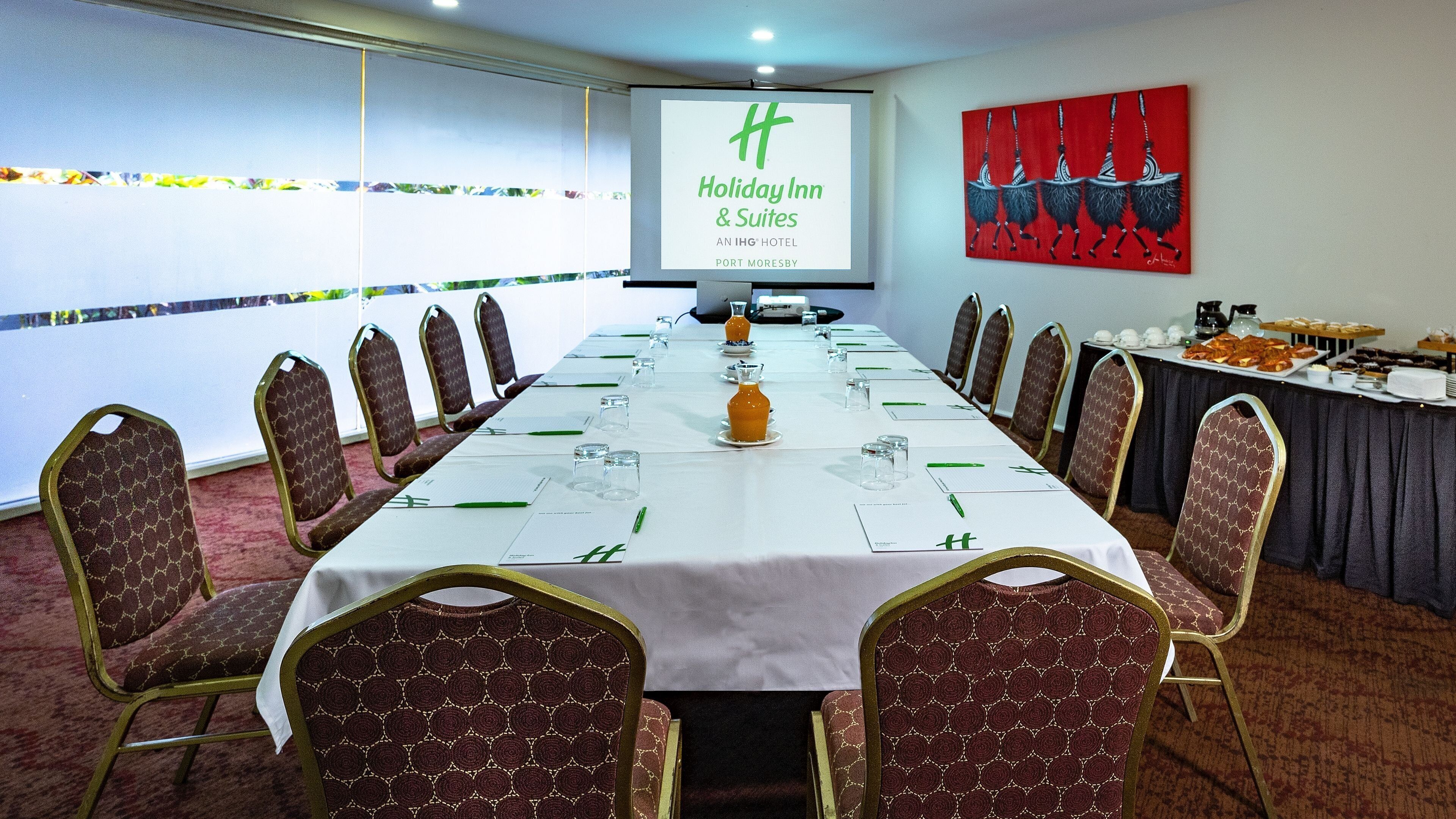 A conference room with a long table, chairs, a projector screen, and a painting on the wall.