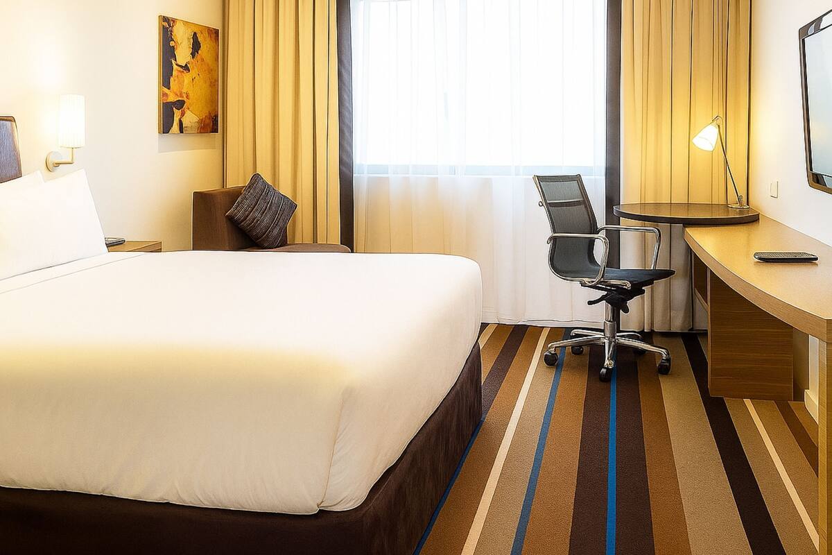 Habitación de hotel con una cama grande, un escritorio, una silla, una lámpara y un cuadro en la pared.