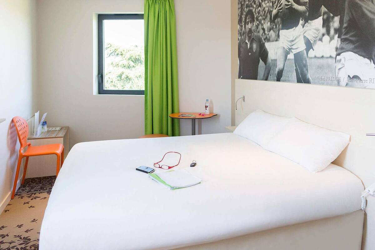 A hotel room with a bed, a desk, a chair, a window with green curtains, and a wall-mounted photo.