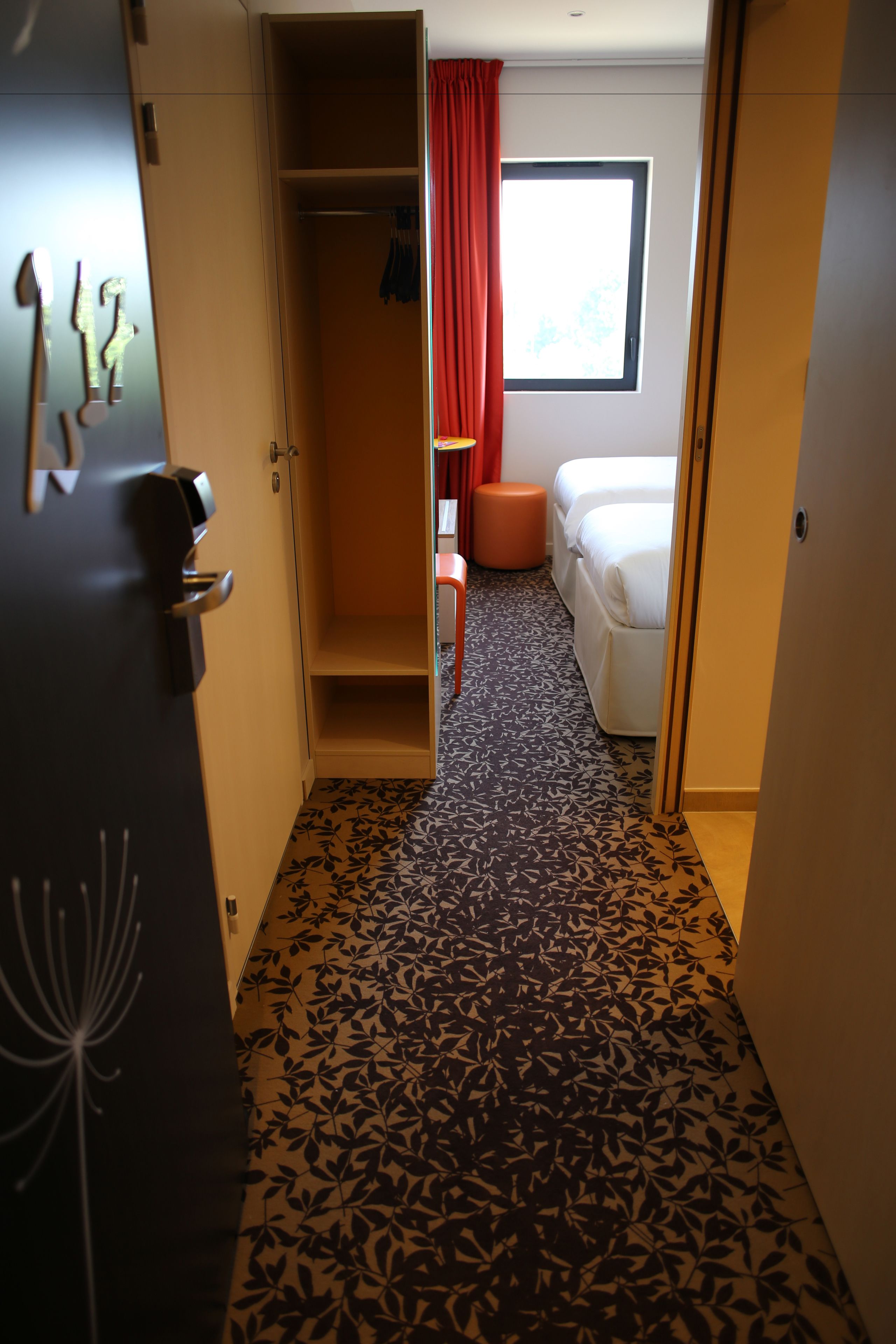 Et hotelværelse med et skab, en seng, et vindue med røde gardiner og et mønstret tæppe.