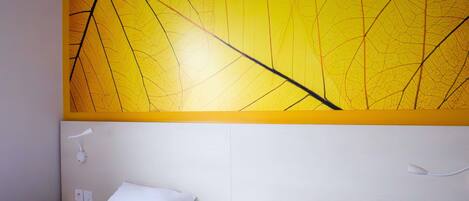Una habitación de hotel con cama, almohadas blancas, una mesita de noche y una lámpara fijada a la pared.