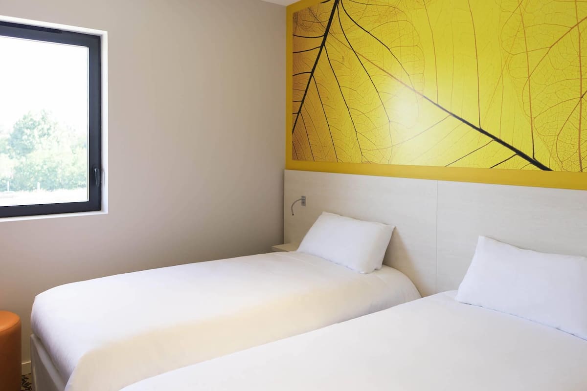 Une chambre d’hôtel avec deux lits, une fenêtre et un mur jaune orné d’un motif de feuilles.